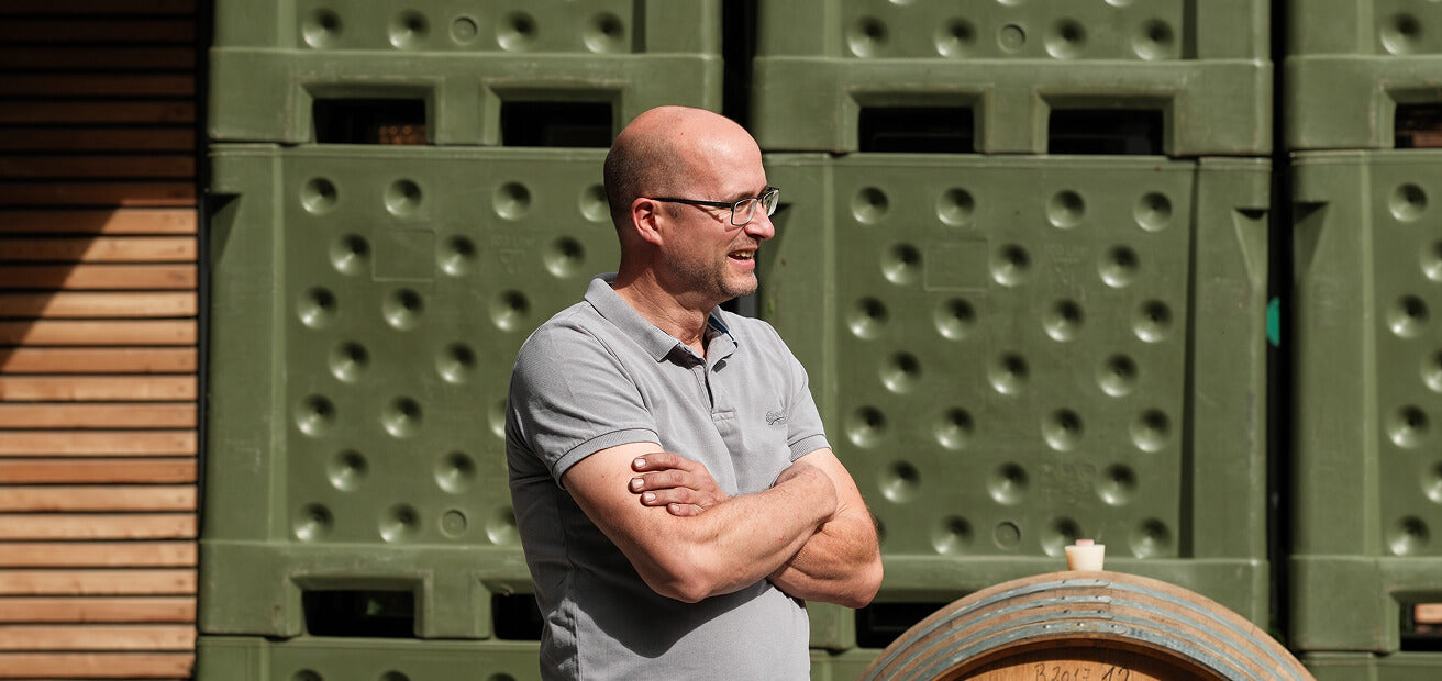 Winzer Johannes Geil-Bierschenk draußen im Weingut, mit verschränkten Armen, lächelnd von der Seite fotografiert